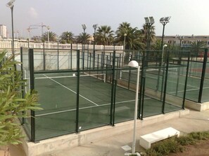 Un court de tennis en plein air, doté d’une surface verte, entouré d’une clôture métallique et de palmiers.