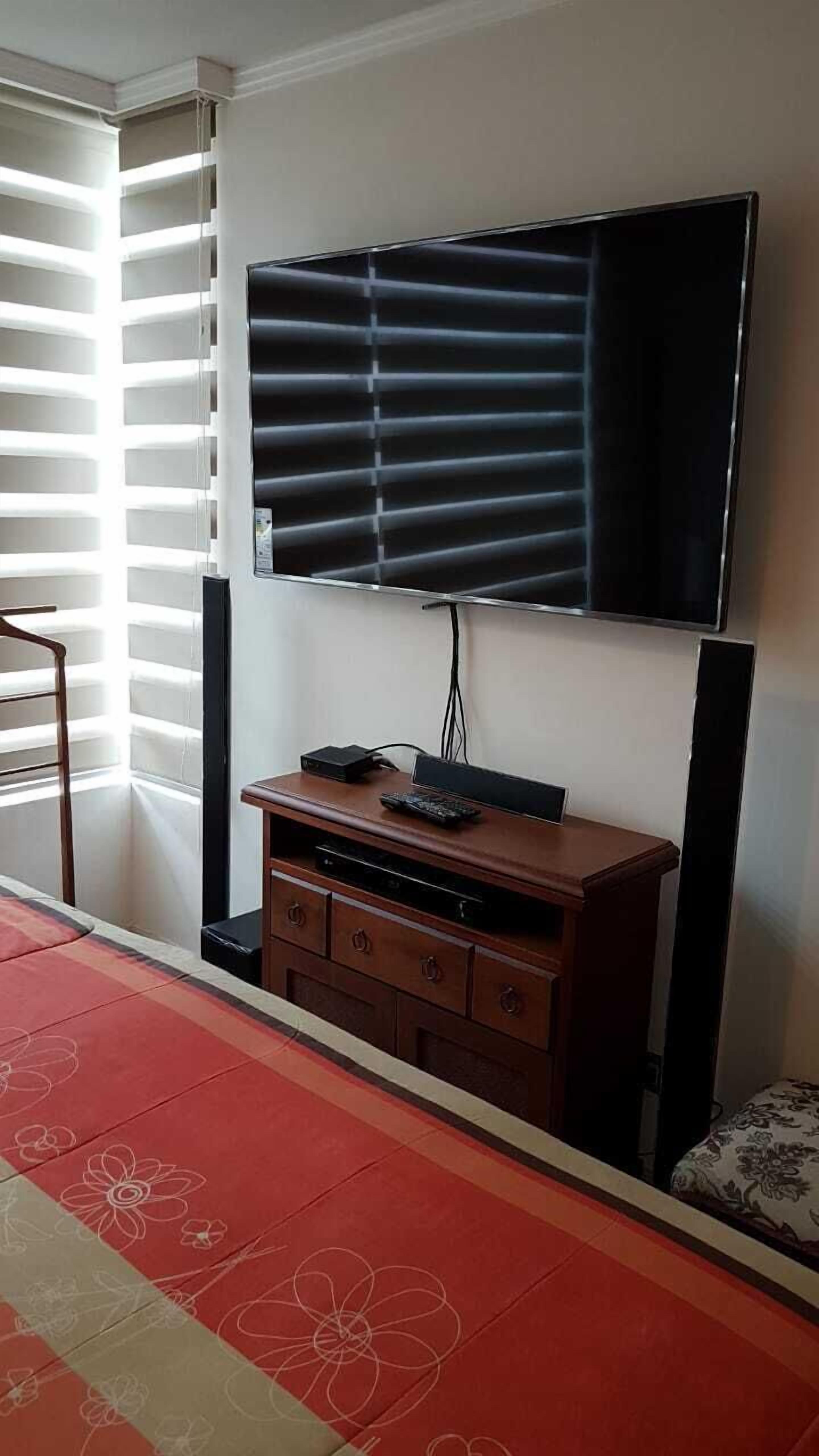 Una habitación con un televisor de pantalla plana grande montado en la pared, un mueble de madera debajo y una alfombra estampada en el suelo.