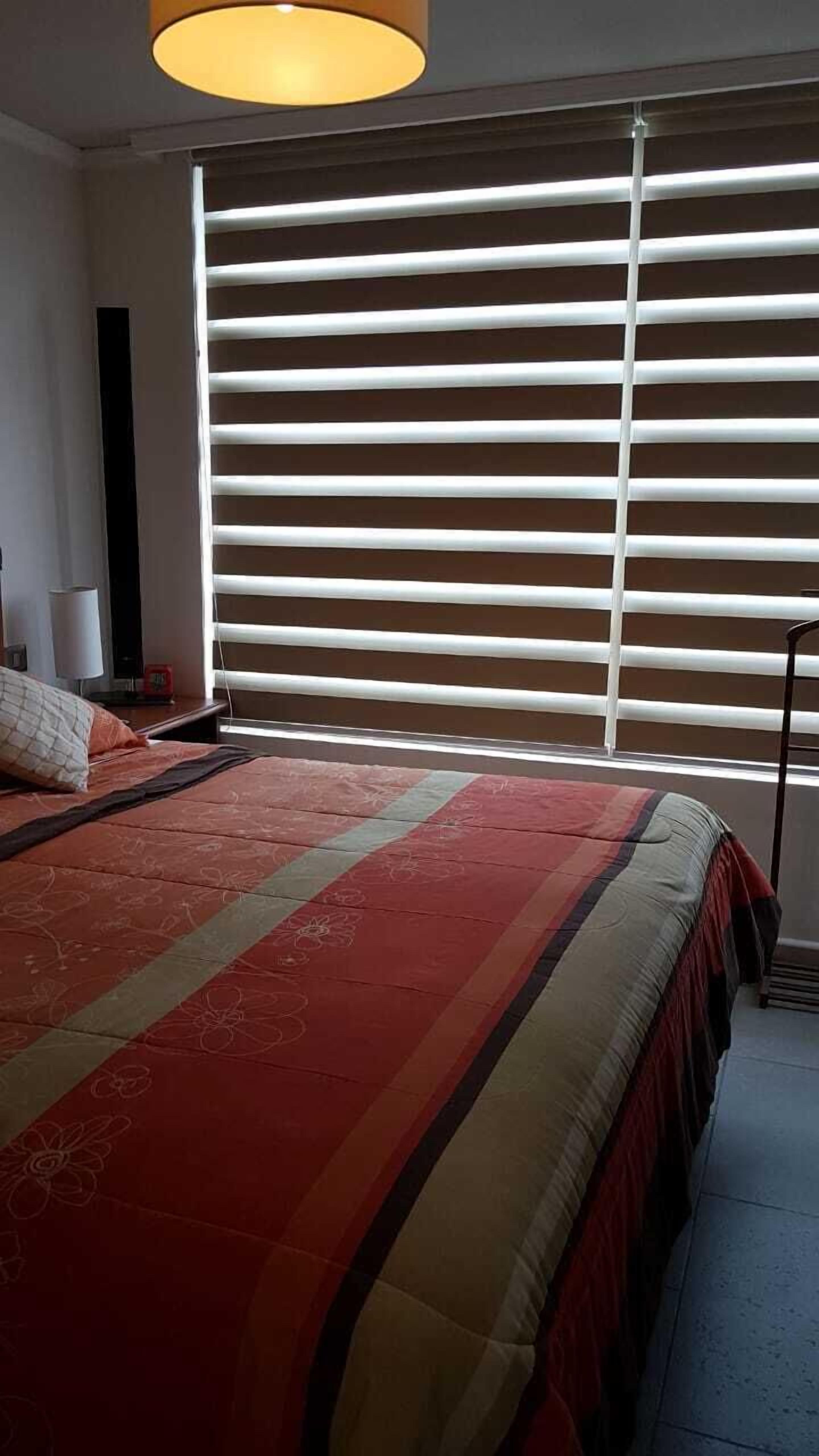 A bedroom with a bed, a bedside table, a lamp, and a window with blinds.
