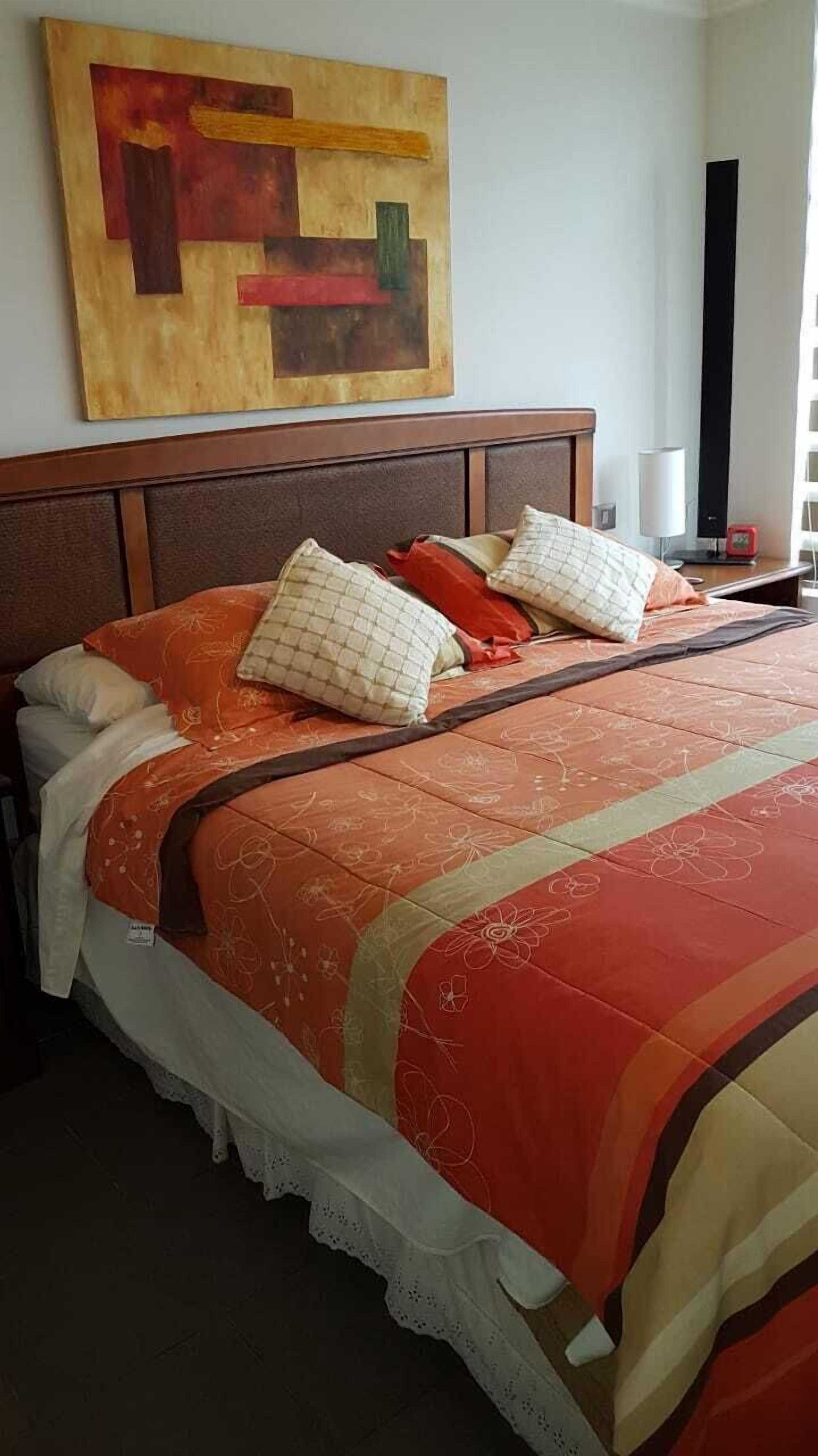 A neatly made bed with a patterned bedspread and pillows, a wooden headboard, a wall art piece, and a bedside table with a lamp and a clock.