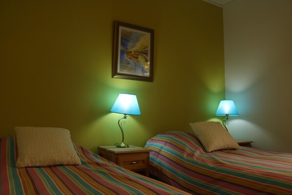A room with two beds, each with striped bedding and a pillow. There are two bedside tables with lamps. A framed picture hangs on the wall above the beds.