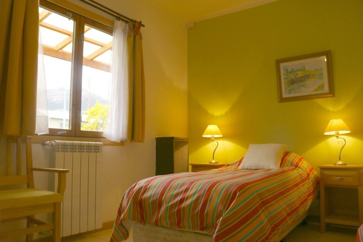 A hotel room with a bed, two bedside lamps, a chair, a radiator, and a window with curtains.
