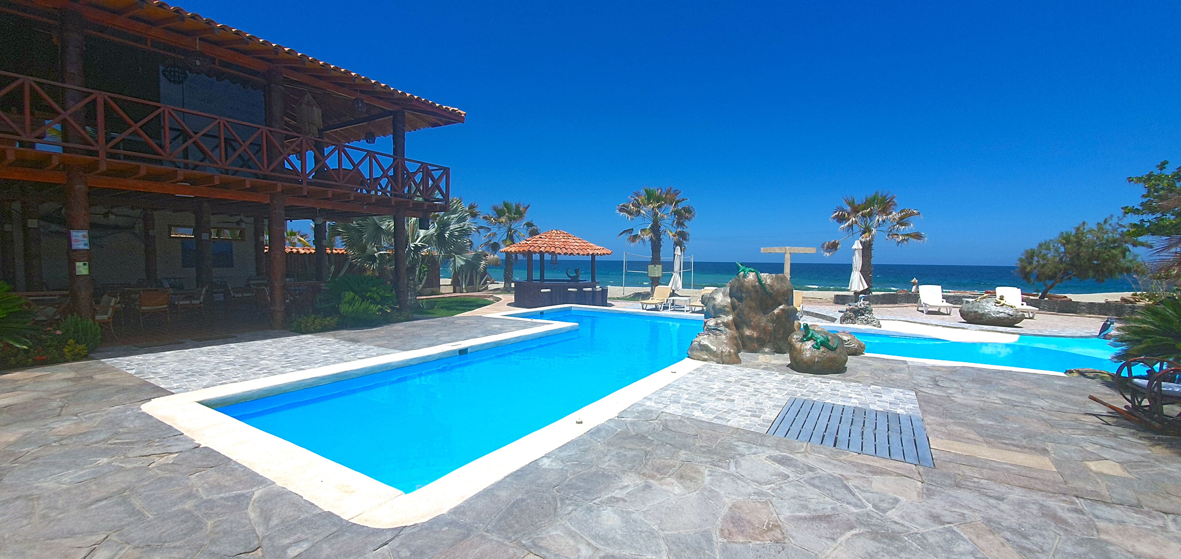 Zona de piscina con vista despejada al océano, terraza de madera y sillones de descanso.