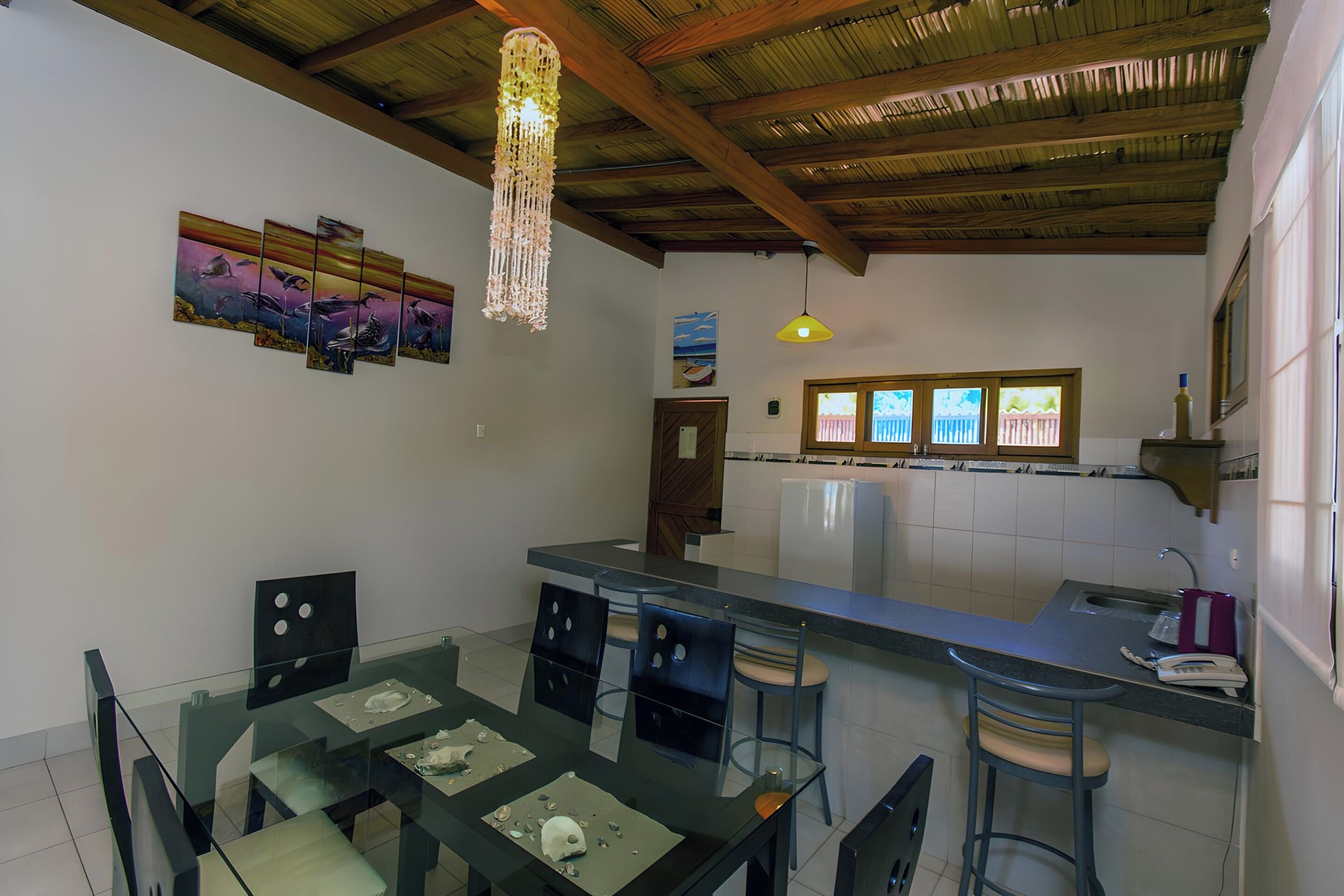 A modern kitchen with a glass dining table, bar stools, a refrigerator, and a hanging light fixture.