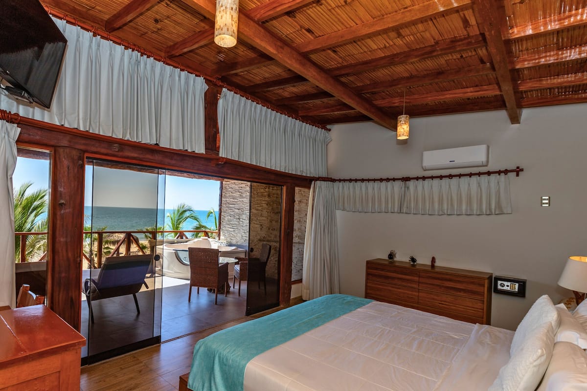 A bedroom with a large bed, wooden ceiling, and a view of the ocean through a sliding door.