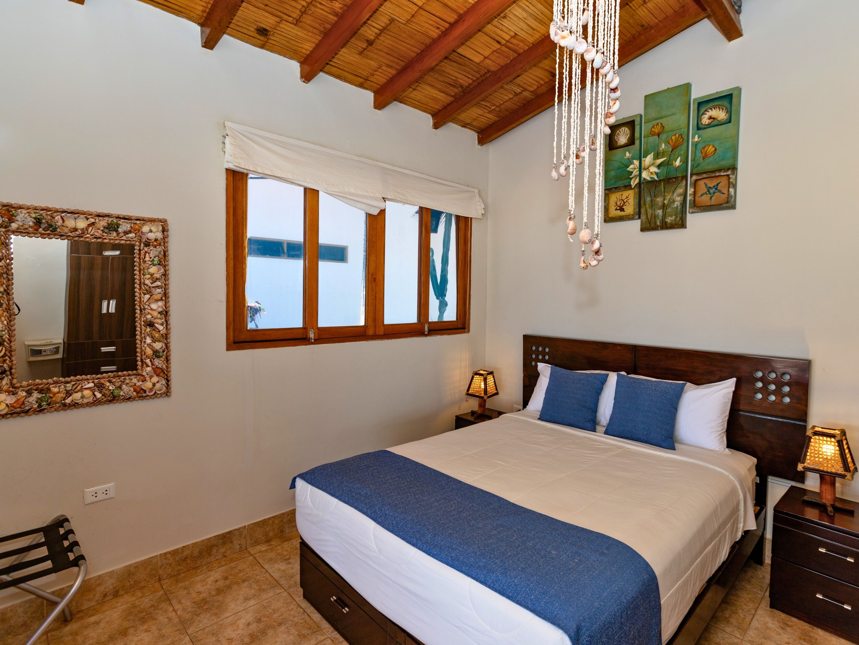 Quarto com uma cama grande, cabeceira de madeira, mesa de cabeceira com abajur, espelho com moldura de concha e uma janela com cortina branca.