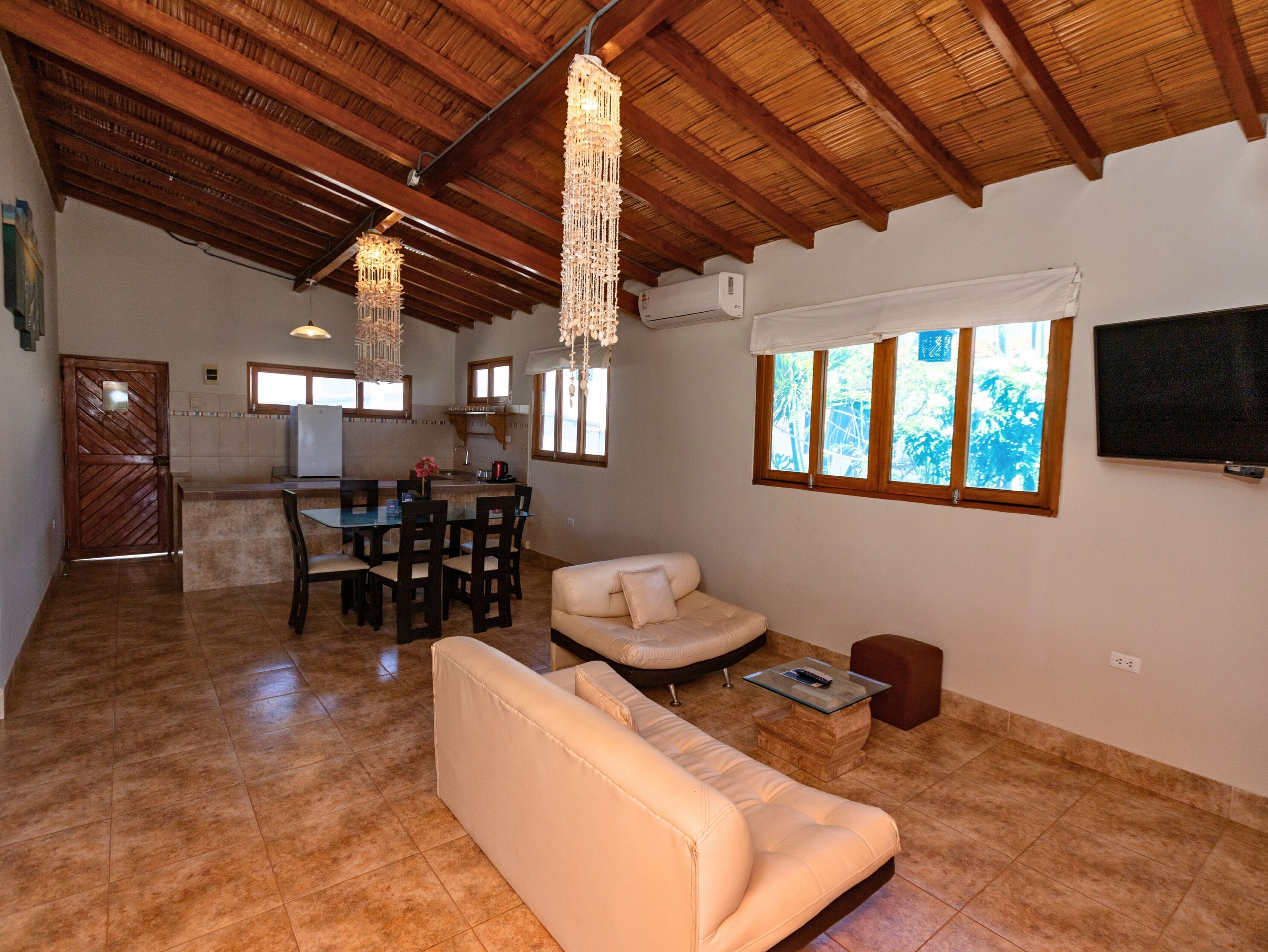 A spacious living area with a wooden ceiling, a dining table with chairs, a sofa, and a TV mounted on the wall.