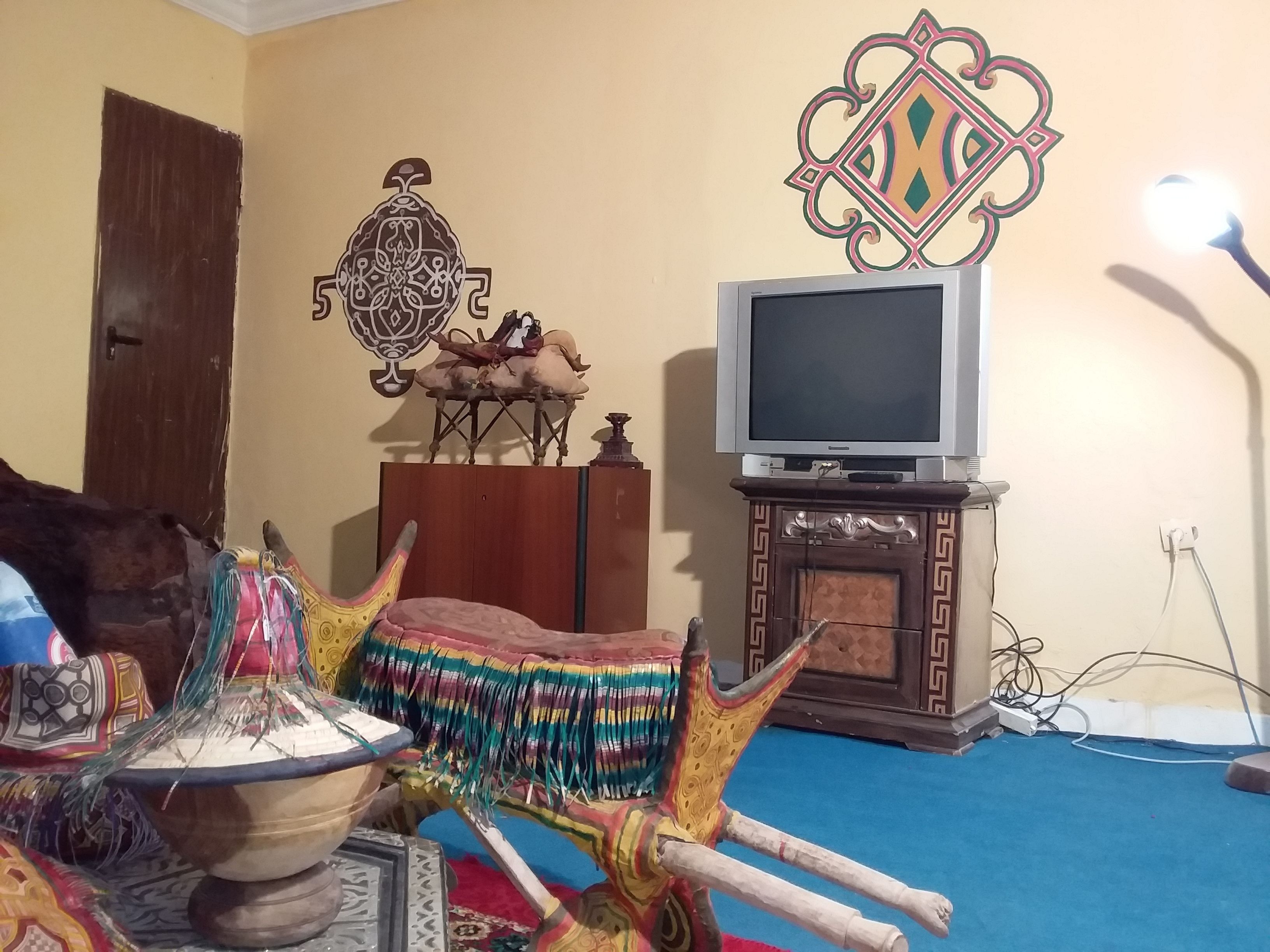 Una habitación con televisión, un adorno en la pared, un mueble de madera y una silla colorida.
