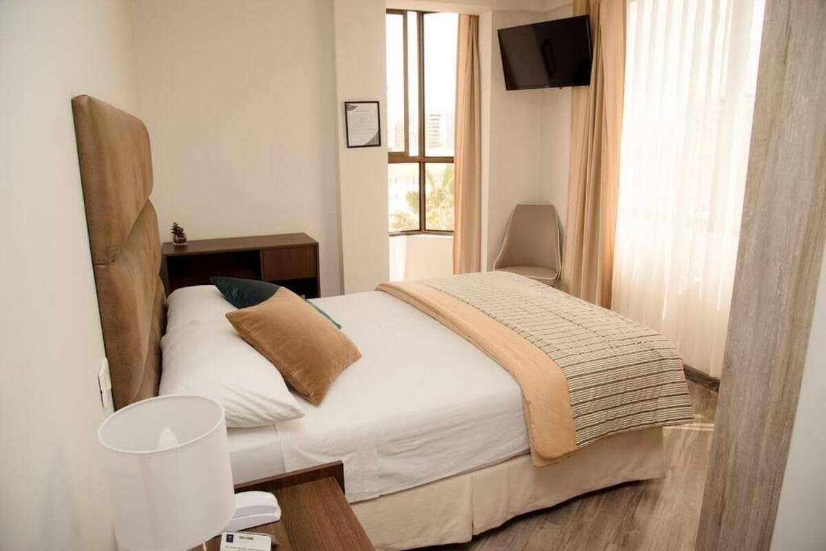 Habitación de hotel con cama, televisión montada en la pared, ventana con cortinas y mesita de noche.