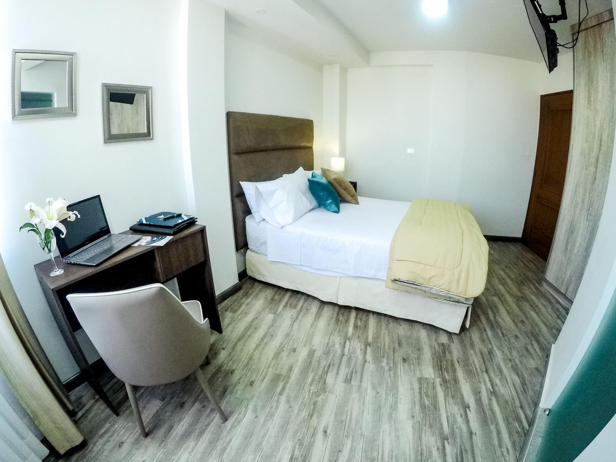 A hotel room with a bed, a desk with a laptop, a chair, and a bedside table.