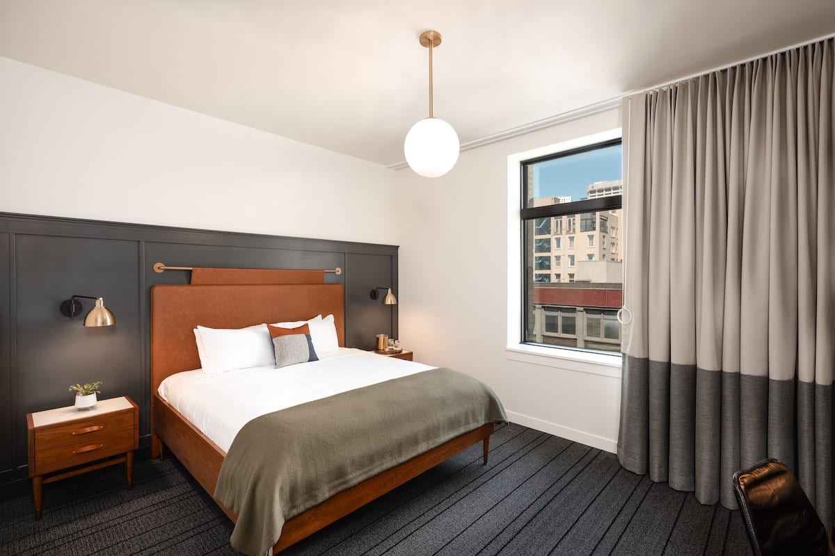 A hotel room with a large bed, a nightstand, a lamp, a window with curtains, and a view of buildings outside.