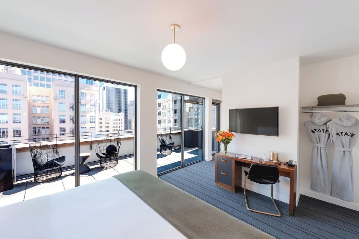 A modern hotel room with a large bed, a desk, a TV, and a balcony with seating.