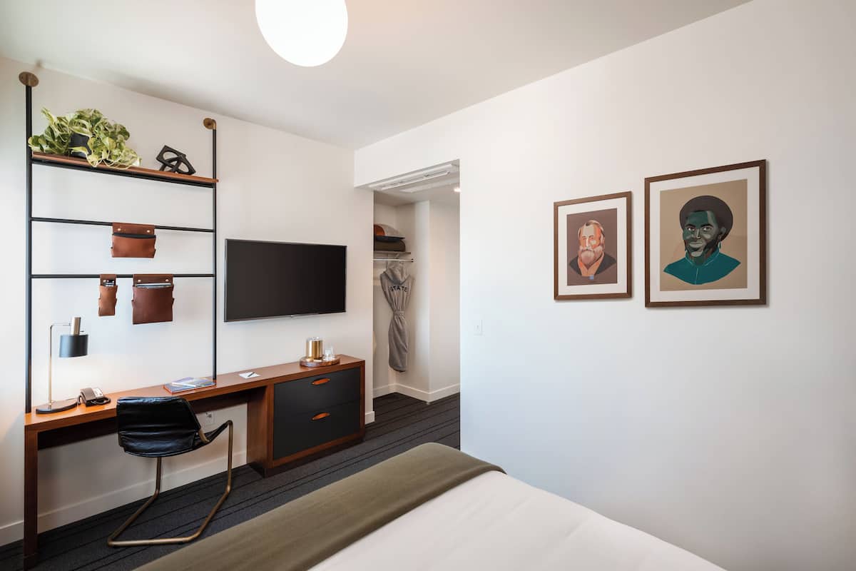 A modern hotel room with a wooden desk, a flat-screen TV, a bed, and two framed artworks on the wall.
