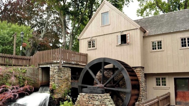 A building with a large water wheel, a stone structure, and a wooden fence.