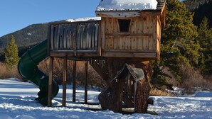 Una casetta gioco in legno con uno scivolo, costruita su un ceppo d'albero, circondata da neve e alberi.