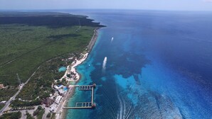 Vista aerea di una zona costiera con mare cristallino, spiaggia sabbiosa e un piccolo resort dotato di piscina e molo.