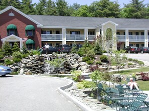 Un hotel con recepción, zona de estar exterior y jardín ajardinado con una pequeña cascada.