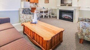 Un salon avec une table basse en bois, des canapés à motifs, une cheminée et une cuisine en arrière-plan.
