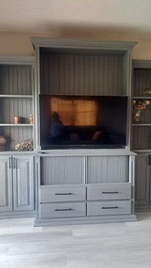 A living room with a large flat-screen TV mounted on a grey cabinet with vertical lines. There are two drawers below the TV and a shelf to the left.