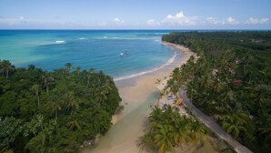An aerial view of a beach with a clear sky, surrounded by dense tropical vegetation.