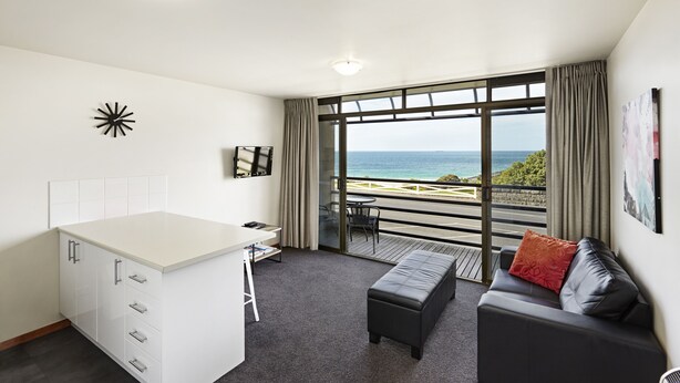 Een moderne hotelkamer met een groot raam met uitzicht op zee, een kleine kitchenette, een bank met kussens en een bureau met een stoel.