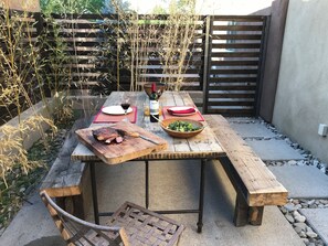 An outdoor dining area with a wooden table, a bowl of salad, a bottle of wine, and a steak on a cutting board. - Truly unique art-filled home w/ garden close to the Plaza. (SANTA FE)