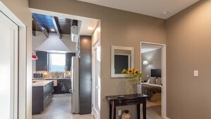 A modern kitchen with dark cabinetry and a microwave, a hallway with a dark floor, a small table with a vase of sunflowers, and a bedroom with a bed and a nightstand.