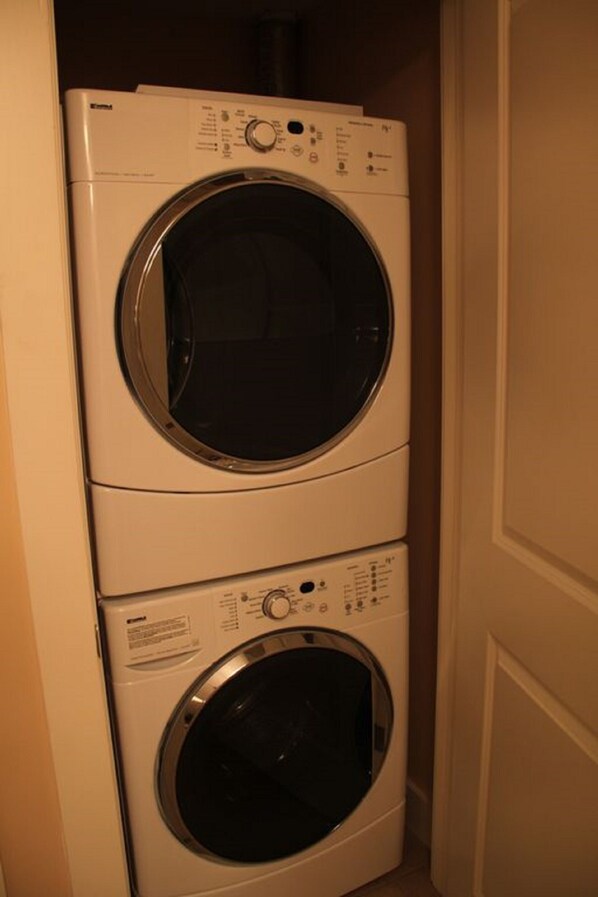 A stacked washer and dryer unit in a room with a door on the right. - Gorgeous, 3 private suites. Balcony View, Pool, Fitness Center! (Princeville)