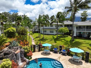 A pool area with a waterfall, outdoor seating, and a view of a two-story building with a balcony.