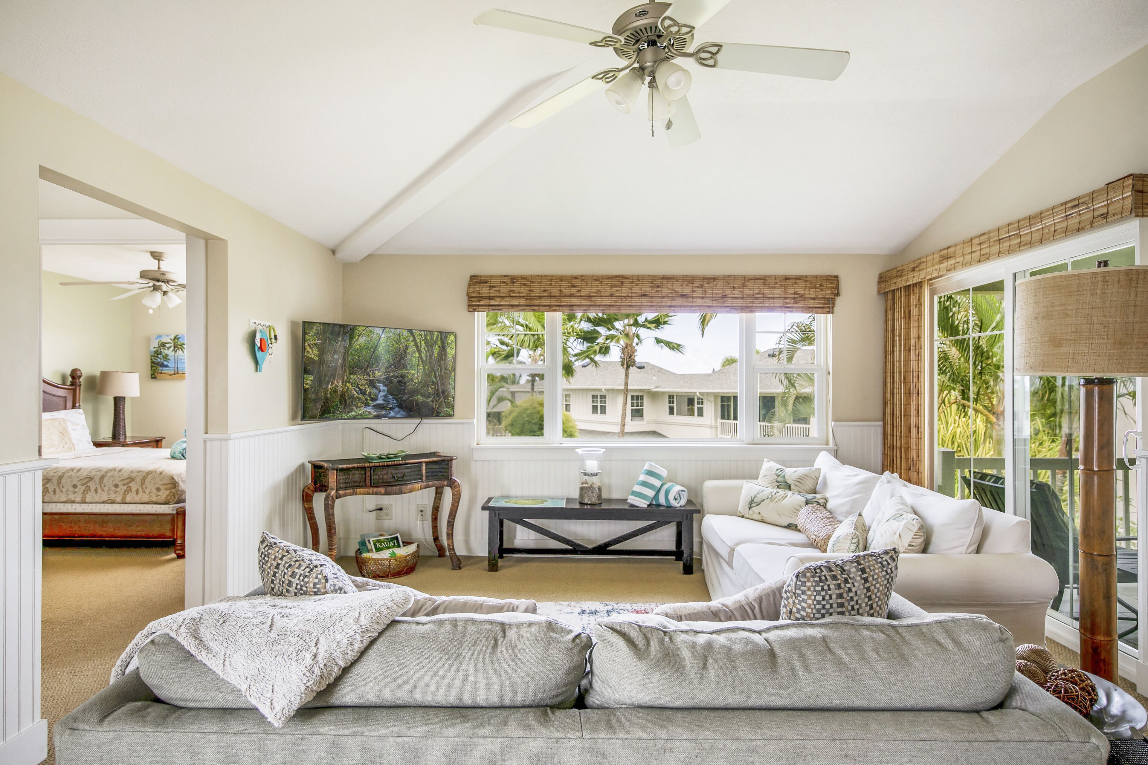 A spacious living room with a large sofa, a coffee table, and a ceiling fan. There is a large window with a view of palm trees and a balcony.