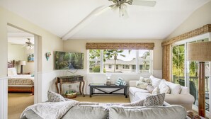 A spacious living room with a large sofa, a coffee table, and a ceiling fan. There is a large window with a view of palm trees and a balcony. - Gorgeous, 3 private suites. Balcony View, Pool, Fitness Center! (Princeville)