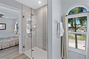 A modern bedroom with a glass-enclosed shower, a bed with floral bedding, and a view of a marina through a window.