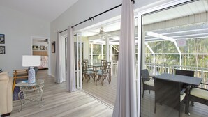 A spacious room with a large sliding glass door leading to an outdoor patio area. There is a dining table with chairs, a small round table with a lamp, and a bed with a headboard. - Hidden Treasure is ready for your stay (Fort Myers Beach)