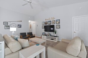 Uma sala de estar com televisão de tela plana, ventilador de teto e parede decorada com quadros.