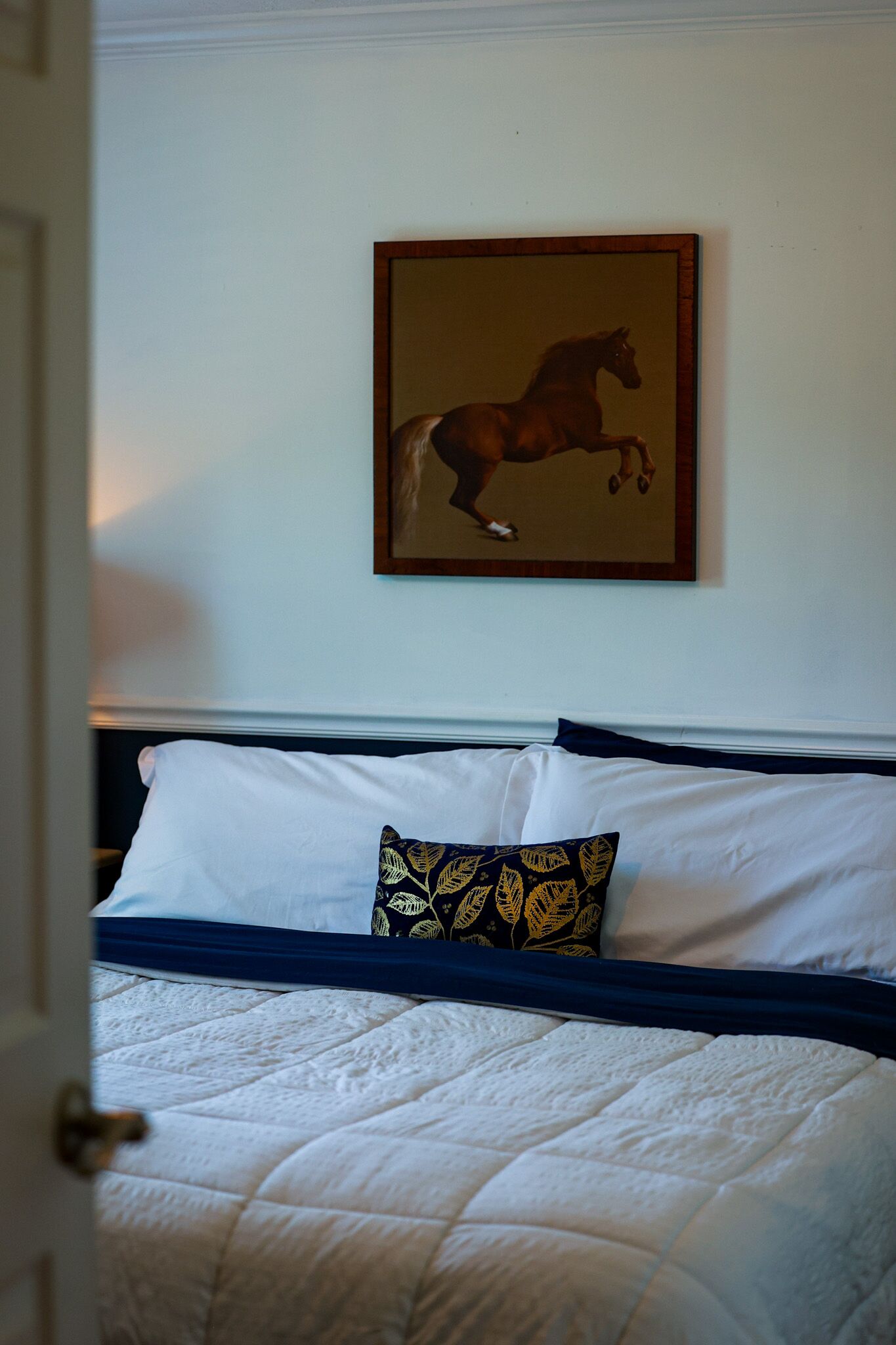A bedroom with a bed, white bedding, a dark blue bedspread, and a framed picture of a horse on the wall.