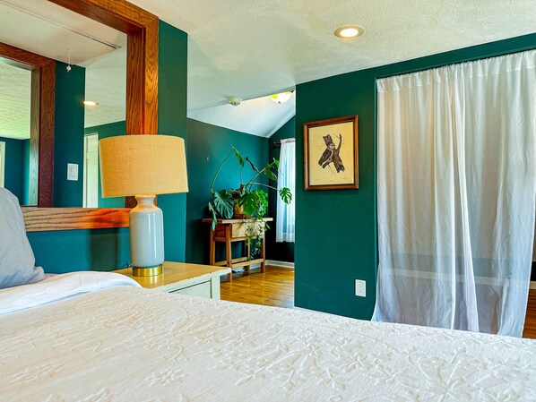 A bedroom with a bed, a bedside table with a lamp, a mirror, a framed picture on the wall, and a window with curtains.