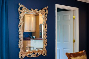 A room with a large ornate mirror reflecting a kitchen area, a white door, and a wooden chair.