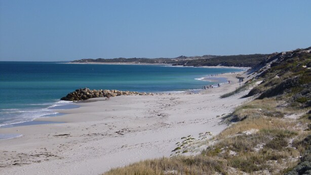 A beach with a rocky outcrop, clear blue water, and a hilly landscape in the background.