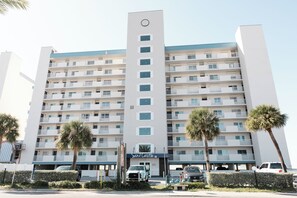 Un hôtel à plusieurs étages, doté d’une entrée centrale, de balcons à chaque étage et de palmiers en premier plan.