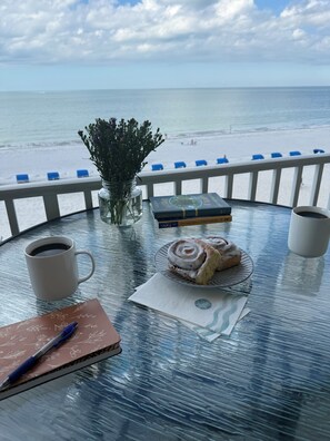Une table avec une tasse de café, un carnet, un stylo, une assiette de pâtisseries, un vase de fleurs et des livres. La table est placée sur un balcon d’où l’on a vue sur la plage et l’océan.