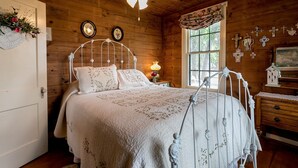 A wooden cabin bedroom with a bed, a ceiling fan, a window with curtains, and wall decorations including crosses and a painting. - Broadway Cottage | Convenient Country Location (Fredericksburg)