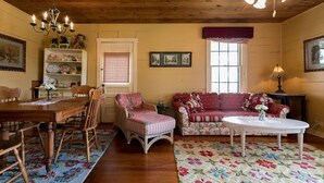 A cozy living room with a wooden ceiling, a dining table with chairs, a floral sofa, a white coffee table, and a bookshelf. - Broadway Cottage | Convenient Country Location (Fredericksburg)