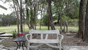 A rustic wooden bench in a wooded area with a small table and a vase of flowers nearby. - Broadway Cottage | Convenient Country Location (Fredericksburg)