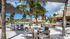 A beachside outdoor seating area with tables and chairs, surrounded by palm trees and a view of a pool and buildings in the background. - ⭐ Blue Bay BEACH villa, minutes walk to beach/pool/golf⭐ 🧡 3BR/2BA (Blue bay golfresort)