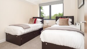 A bedroom with two beds, a window with a patterned curtain, and a bedside table with a lamp.