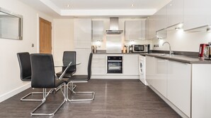 A modern kitchen with white cabinets, a built-in oven, a microwave, and a coffee machine. There are four black chairs around a glass table.