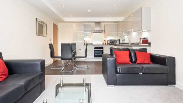 A modern living room with a glass coffee table, black leather sofas with red cushions, a dining area with a chair, and a kitchen with white cabinets and appliances.