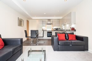 A modern living room with a glass coffee table, black leather sofas with red cushions, a dining area with a chair, and a kitchen with white cabinets and appliances. - 4 Chapel Apartments · Stylish 2-Bed Flat Near York City Centre - Pass the Keys (York)