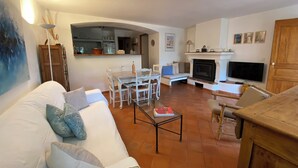A living room with a sofa, coffee table, and TV. There's a fireplace, dining area, and kitchen in the background. - *** Les BOUTINELLES * Superb apartment in LA FERME DU MOULIN ROUX (Ramatuelle)