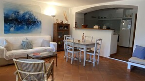 A living room with a white sofa, a dining table with chairs, a wicker chair, and a painting on the wall. - *** Les BOUTINELLES * Superb apartment in LA FERME DU MOULIN ROUX (Ramatuelle)